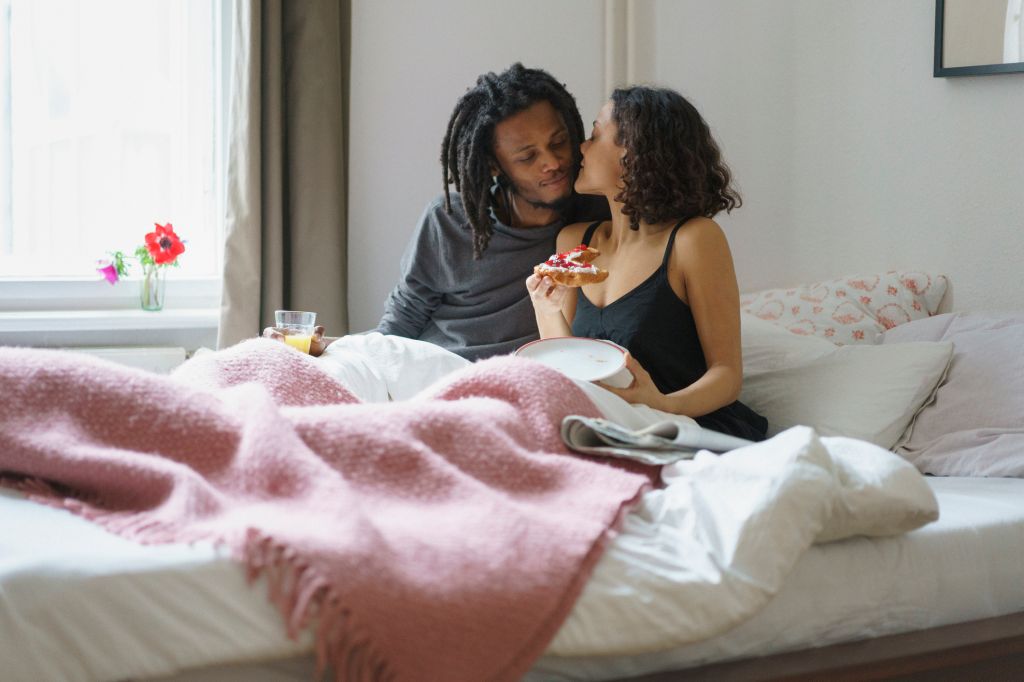 Young woman kissing man while holding pie in bed at home