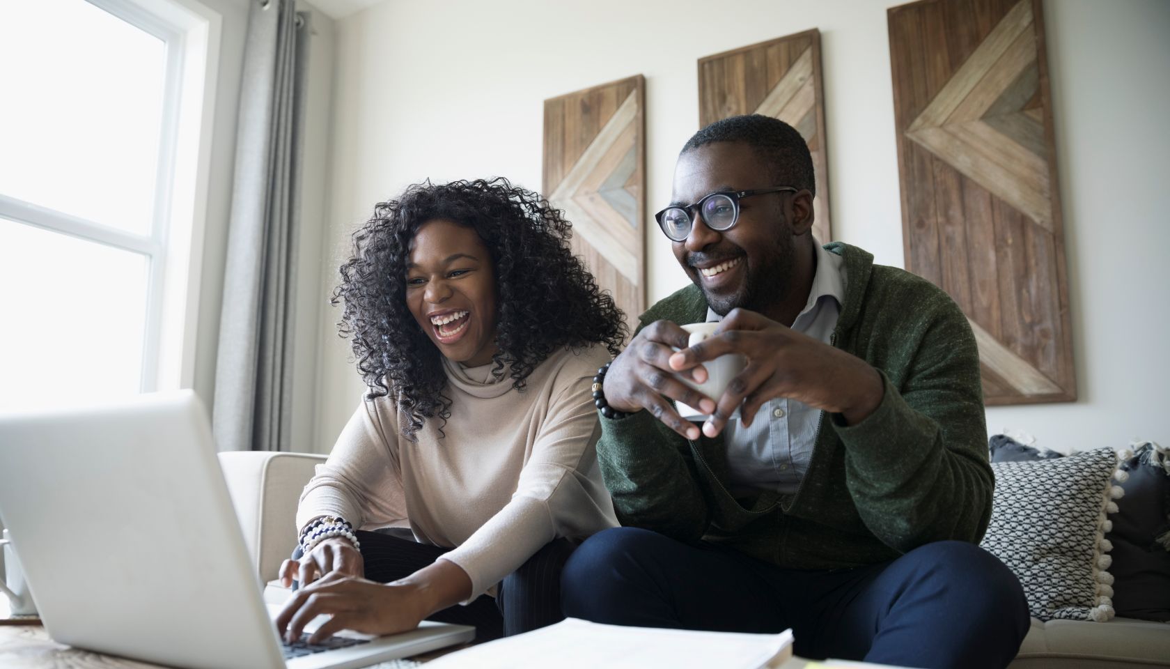 Laughing couple with laptop drinking coffee and paying bills online in living room