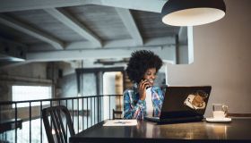 Young woman, African-American Ethnicity, working at laptop in cafe, using mobile phone.