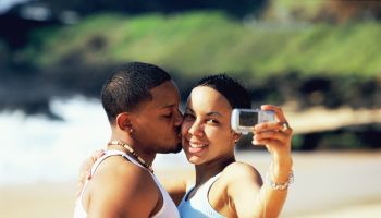 Young man kissing woman using digital camera at beach