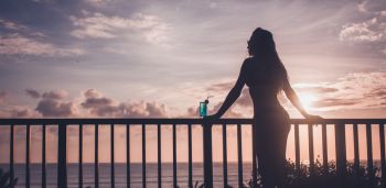 Silhouette of sensual woman drinking blue lagoon cocktail on tropical balcony at sunset.