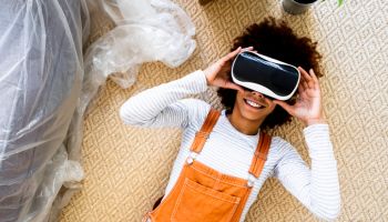 Afro woman wearing virtual reality simulator while lying on carpet in new apartment