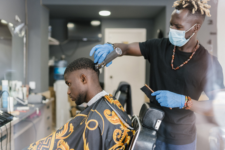 Barber with locs wearing mask and glove while cutting customer's hair in salon