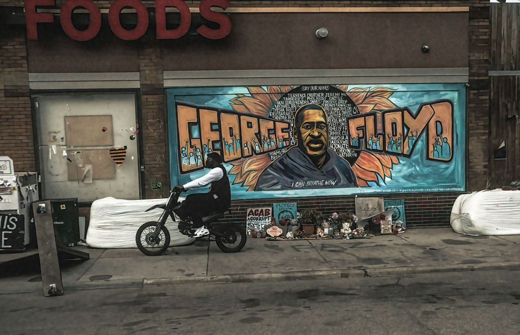 A man rides a motorcycle next to a mural of George Floyd by...