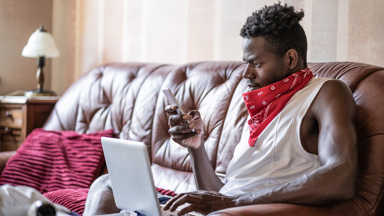 African guy resting on sofa at home with laptop, watching his phone, enjoying lazy free day. Lifestyle concept. Home office. Work from home.