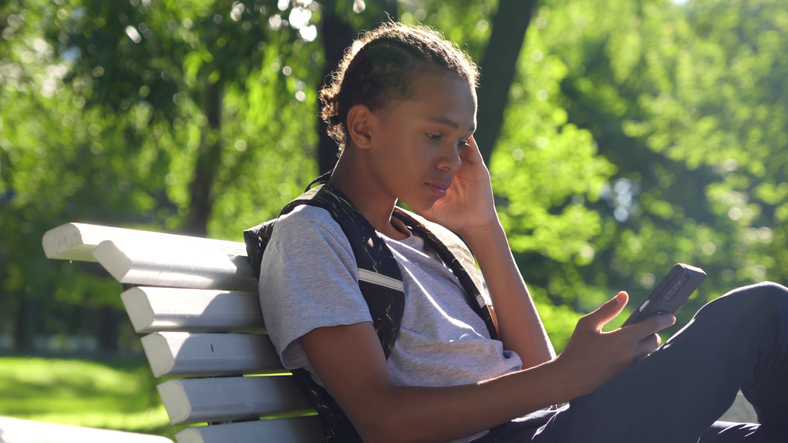 African-American teen boy with smartphone and backpack sit on bench in park.