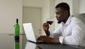 Young African man browsing the internet on a laptop in the kitchen and drinking red wine at home