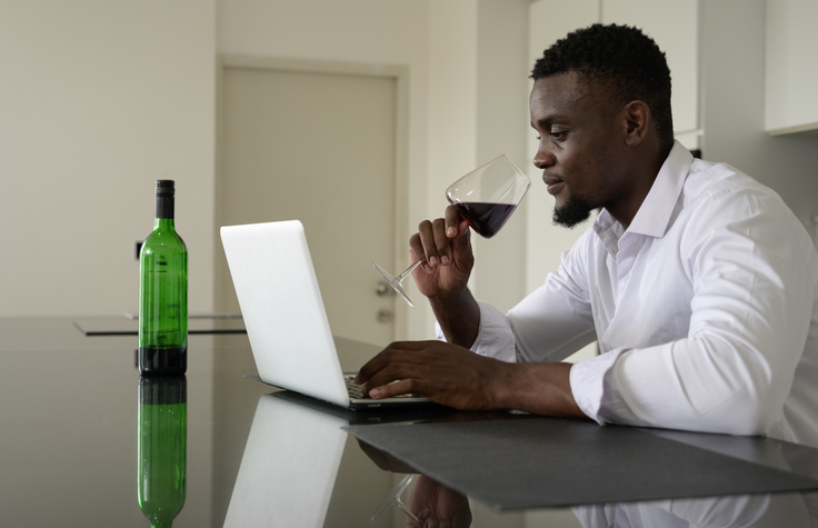 Young African man browsing the internet on a laptop in the kitchen and drinking red wine at home