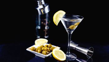 Glass of dry martini cocktail with olives and lemon with plate of olives on a black background dark photography