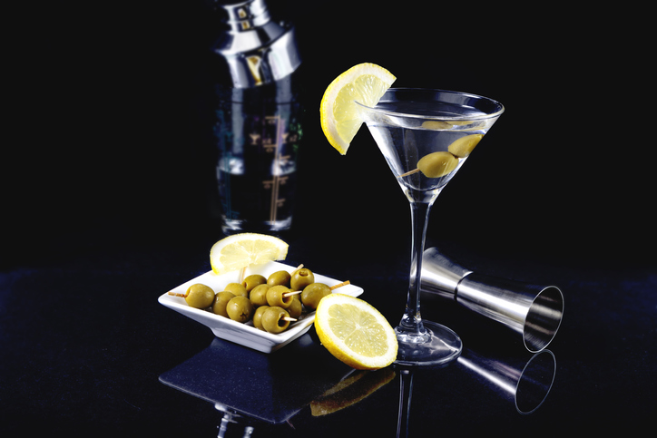 Glass of dry martini cocktail with olives and lemon with plate of olives on a black background dark photography