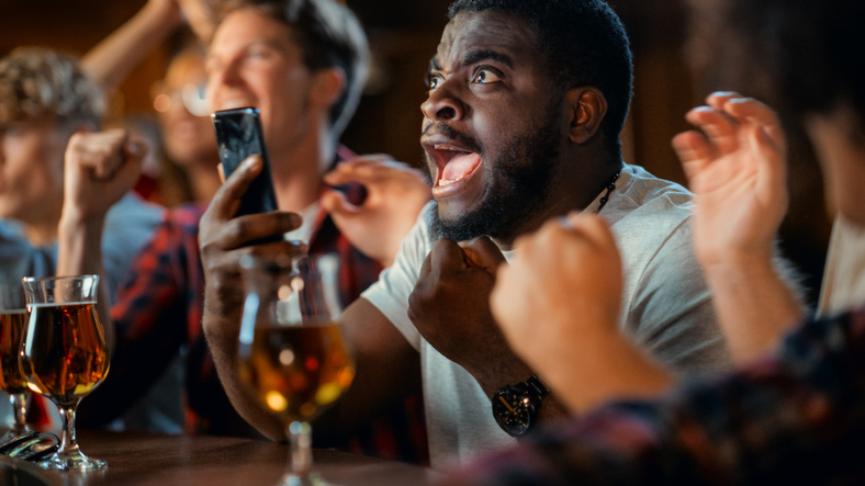 Portrait of an Excited Young Black Man Holding a Smartphone, Stressed About a Sports Bet on His Favorite Soccer Team. Lively Successful Emotions When Football Team Scores a Winning Goal.