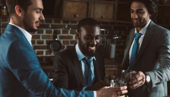 smiling young multiethnic male friends in suits clinking glasses of whiskey