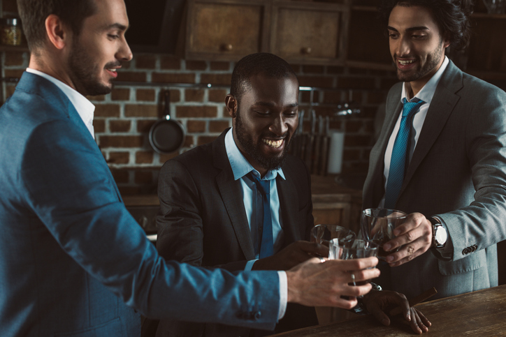 smiling young multiethnic male friends in suits clinking glasses of whiskey