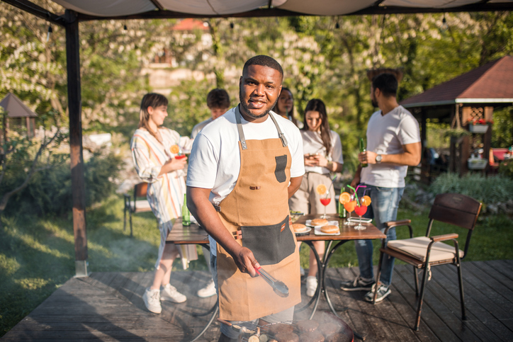 A black man with an apron prepares a barbecue for his friends. They are in the garden and enjoying each other's company