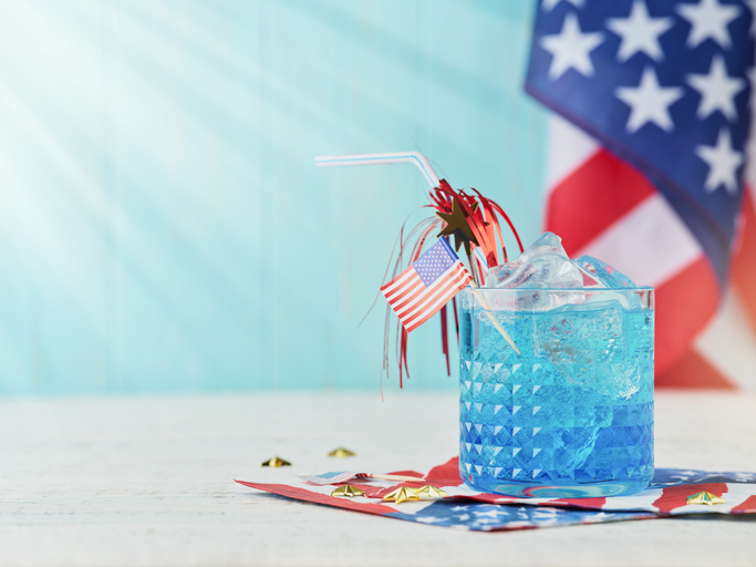 4th of July Holiday background. Cocktail with blue liqueur, American flag and festive straw against background of old turquoise board and sun rays. Patriotic American Flag drinks. Mock up.