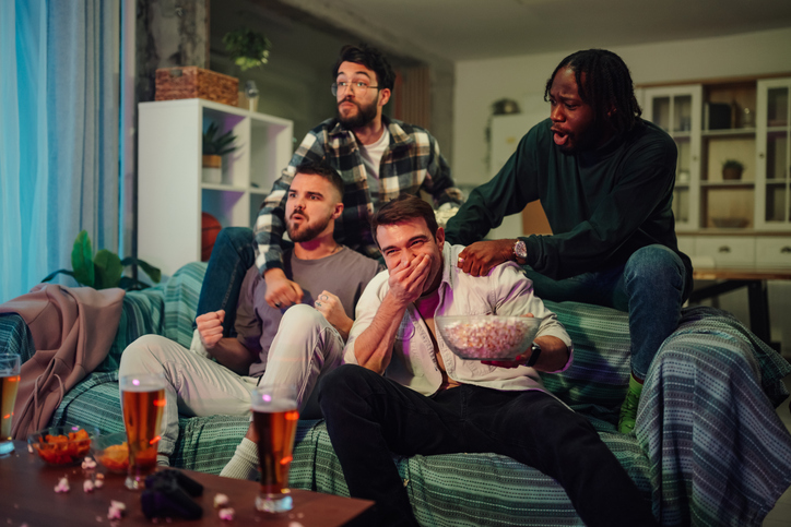 Diverse male friends watching exciting sports on tv at home, cheering and reacting emotionally while relaxing on sofa with snacks