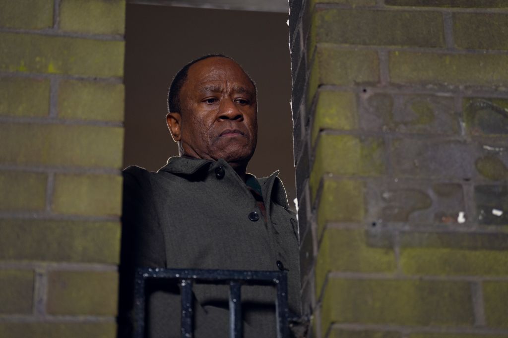 A middle-aged African American man with a serious expression standing in a dimly lit hallway against a brick wall.