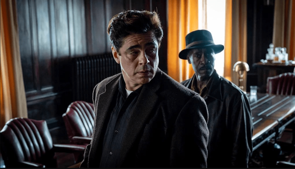 Two men in dark coats standing in a dimly lit room with orange curtains in the background.