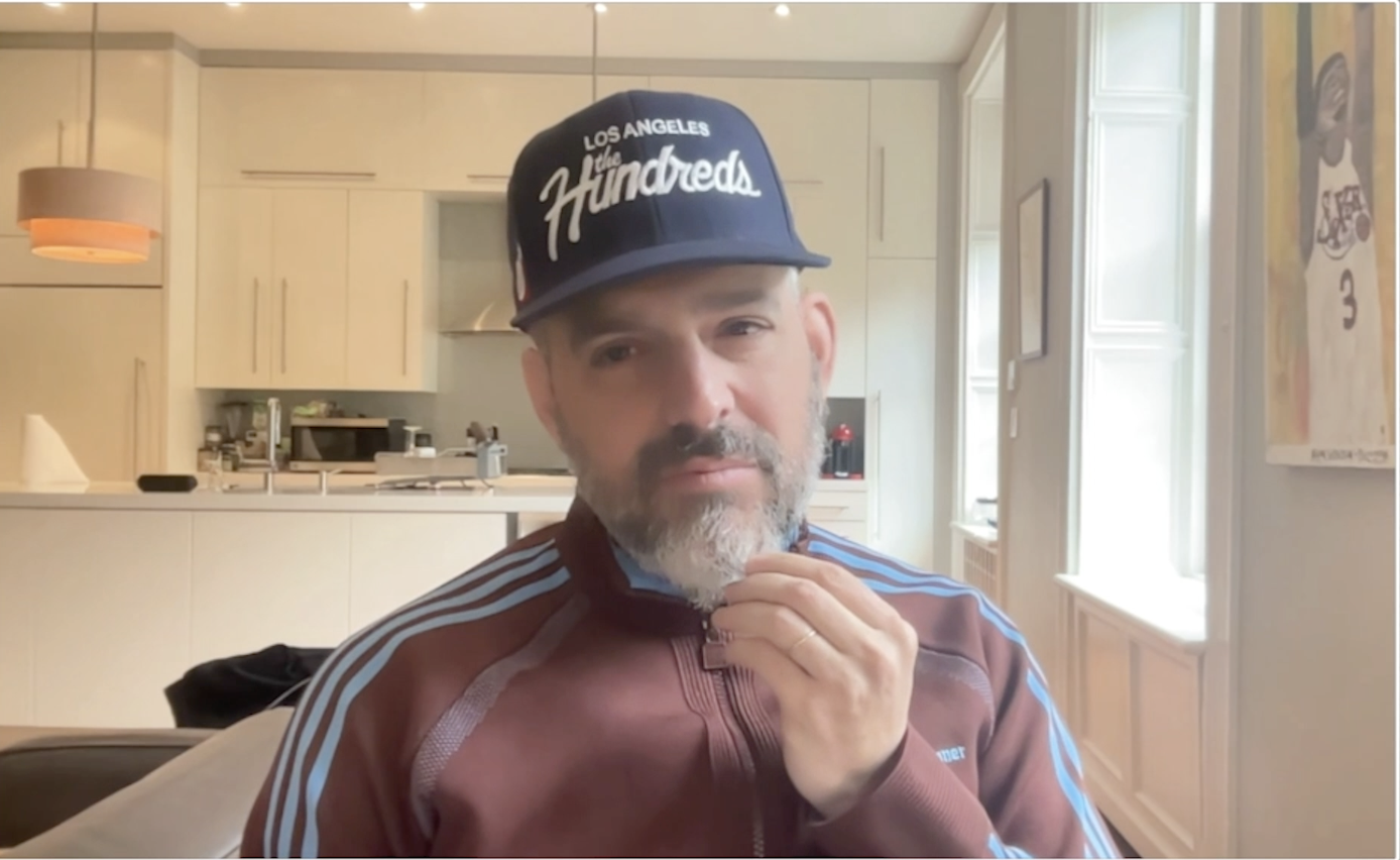 A bearded man wearing a Los Angeles Hundreds baseball cap and a blue and white striped jacket, standing in a kitchen.