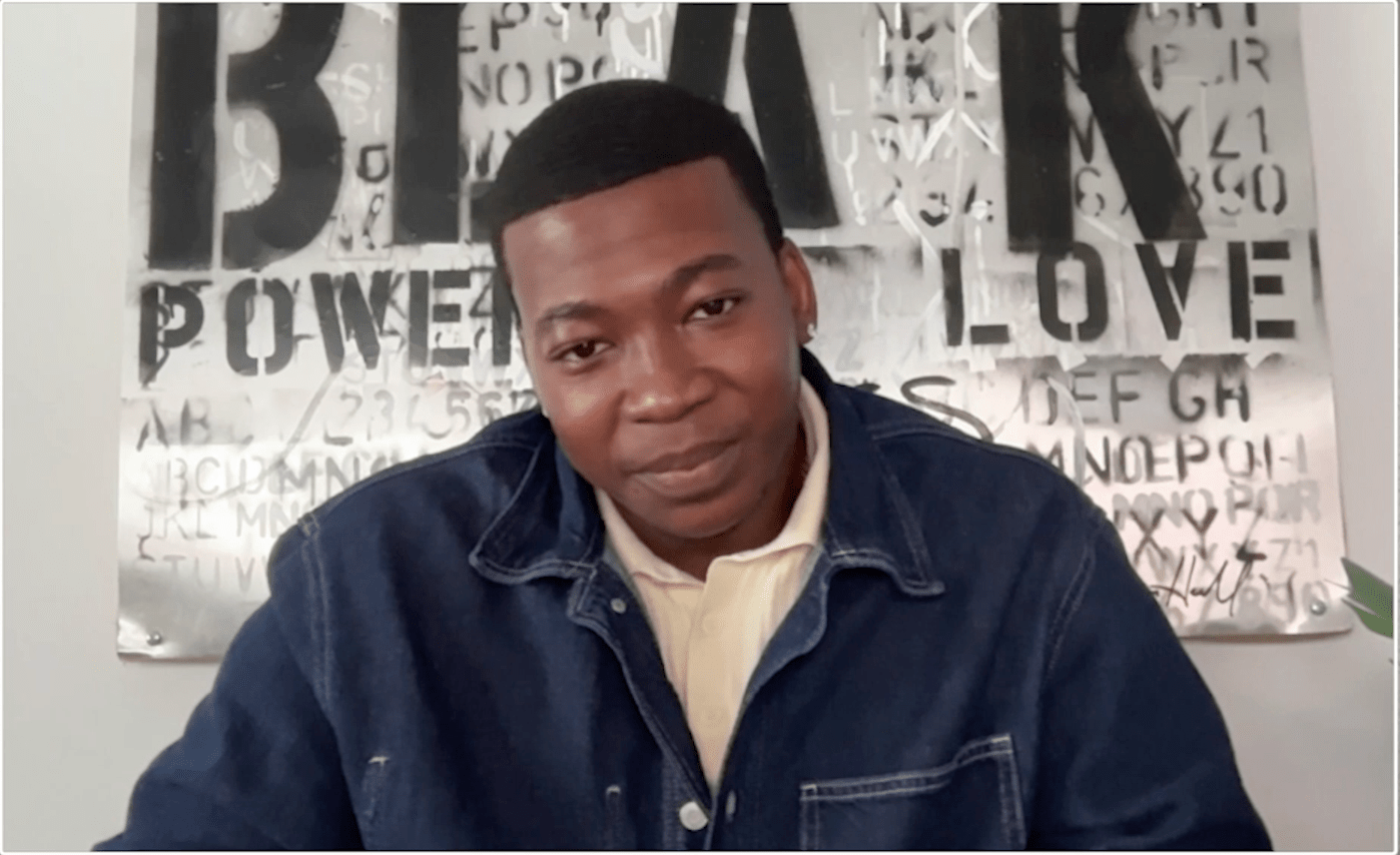 A Black man wearing a navy blue jacket and a light-colored shirt, sitting in front of a wall with graffiti and text that includes "POWER" and "LOVE".