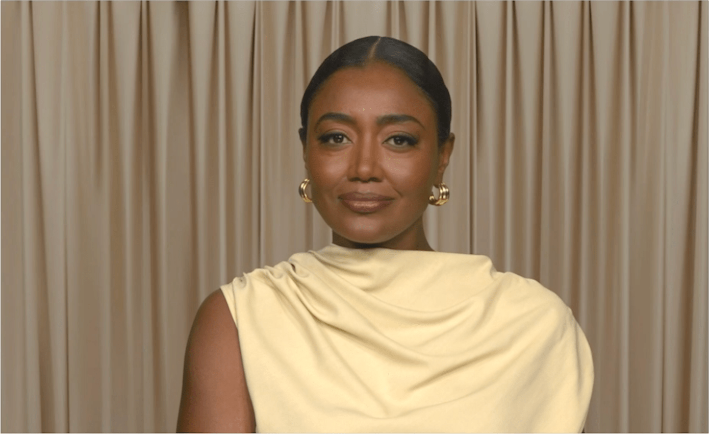 A woman with dark skin and hair wearing a beige dress with a draped neckline, looking directly at the camera with a slight smile.