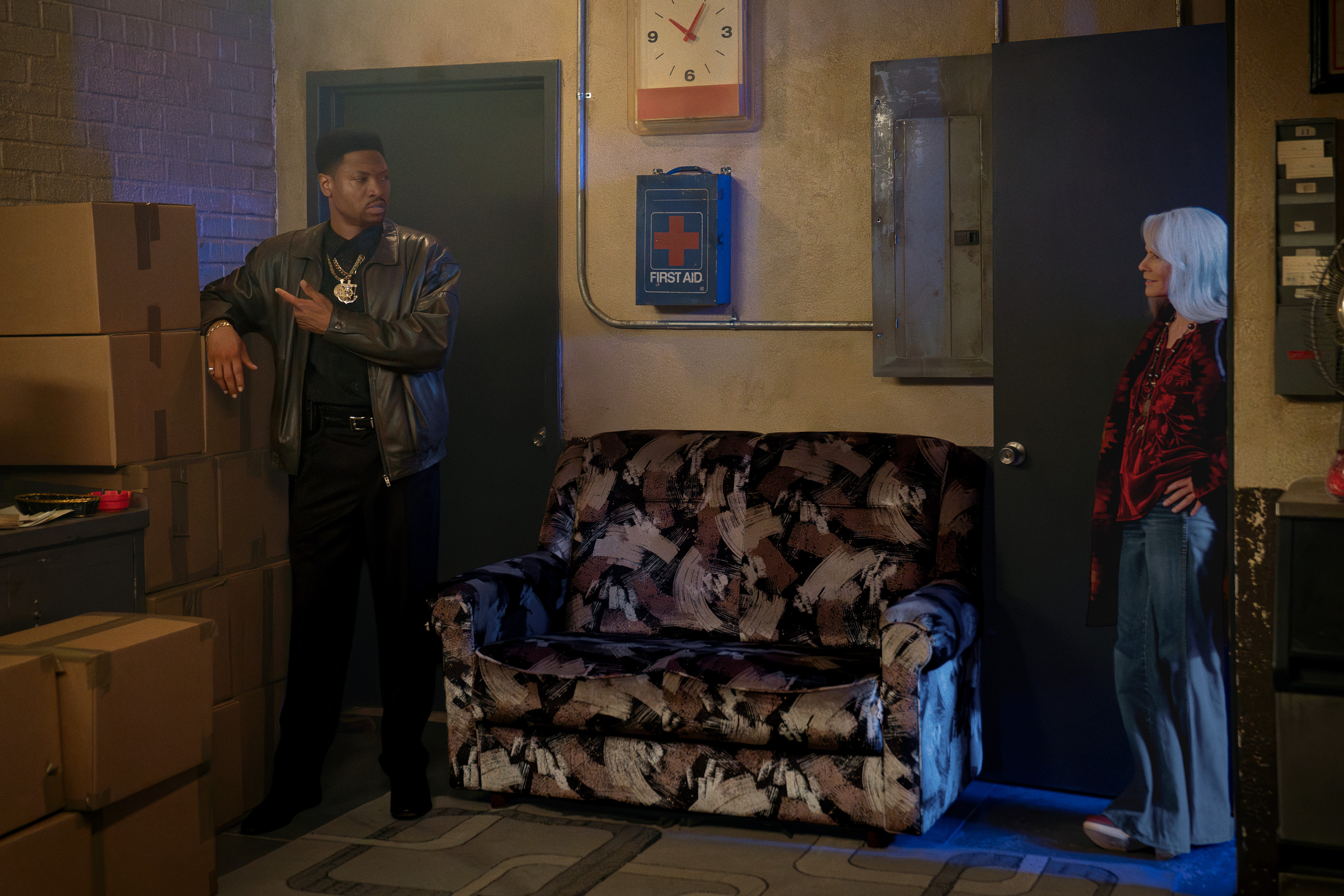 Two people, a man in a leather jacket and a woman in a red floral top, standing in a room with a First Aid kit and a clock on the wall.