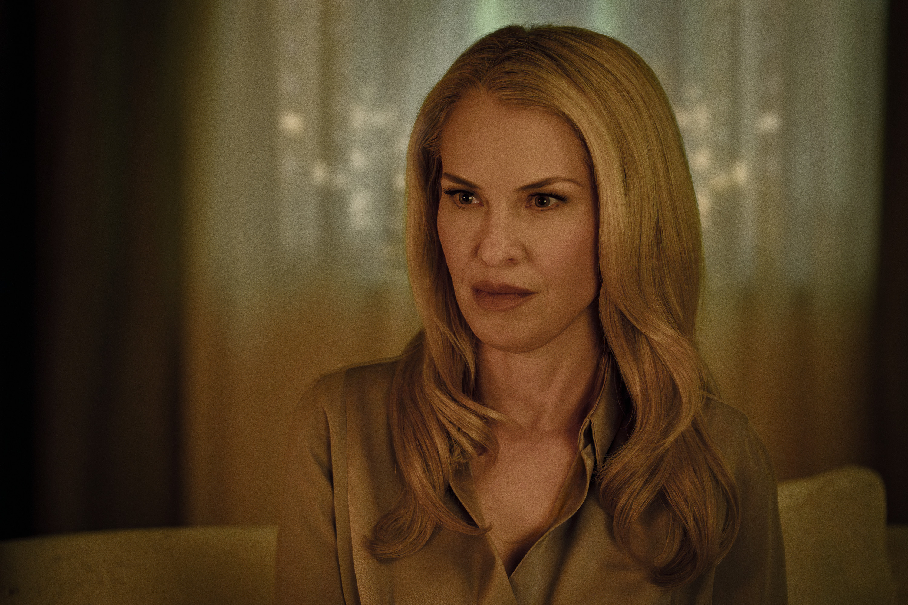 A woman with long blonde hair wearing a tan coat looks directly at the camera with a serious expression.