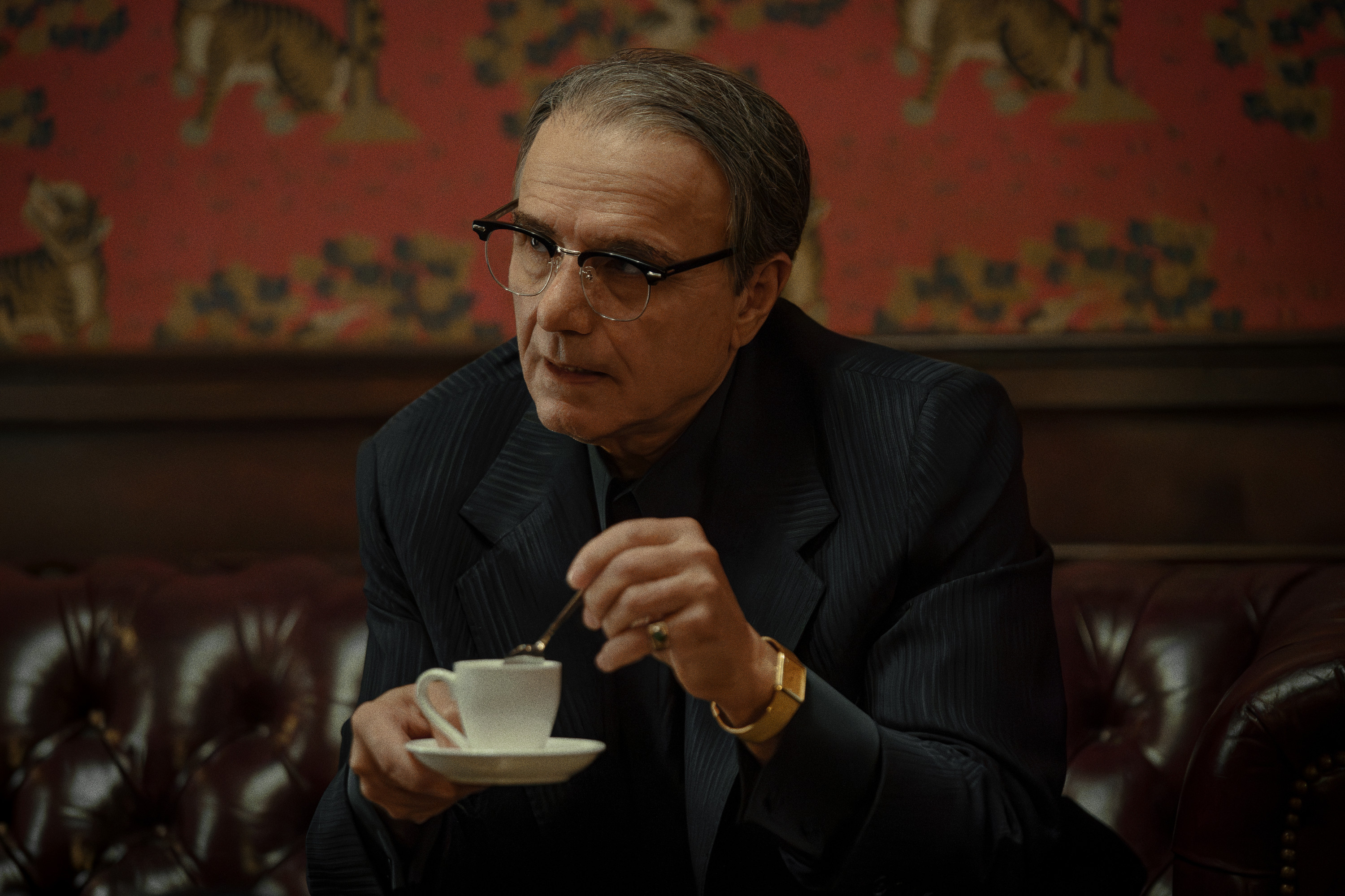 A man in a dark suit and glasses sitting at a table, holding a cup of coffee and looking pensive.