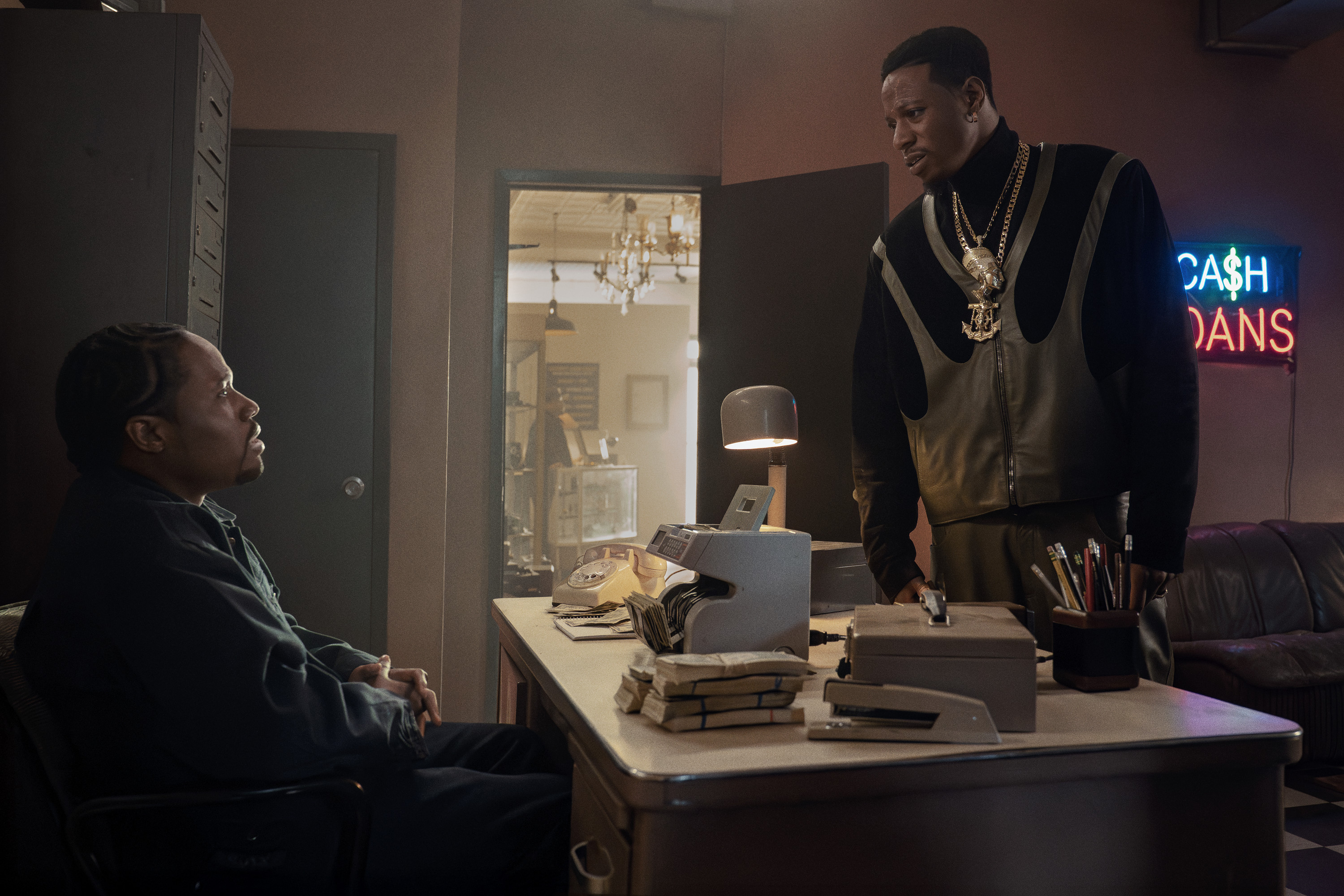 Two men, one sitting at a desk and the other standing, in a room with a neon "Cash Loans" sign.