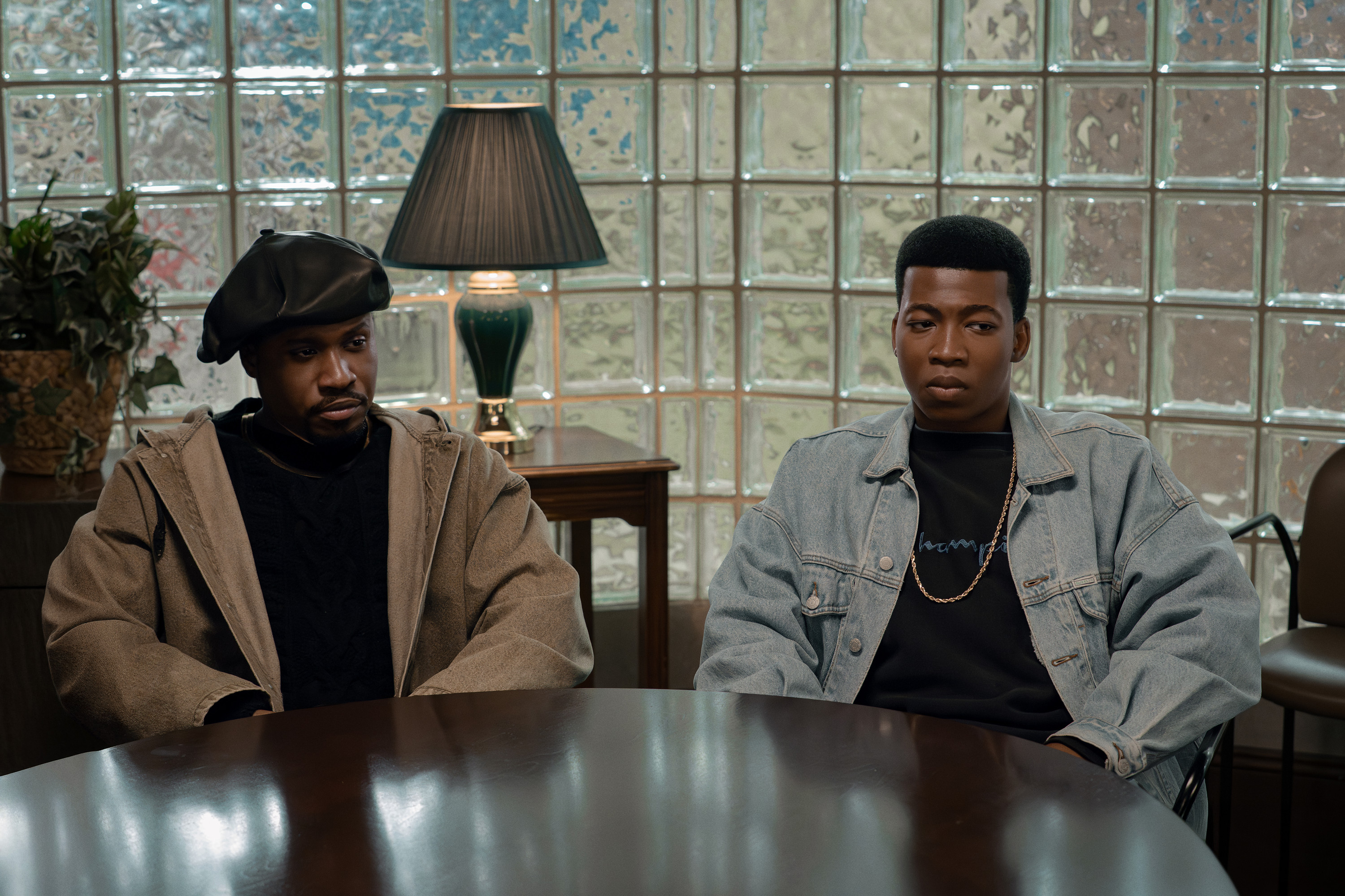 Two Black men sitting at a table in a room with glass block walls and a lamp.