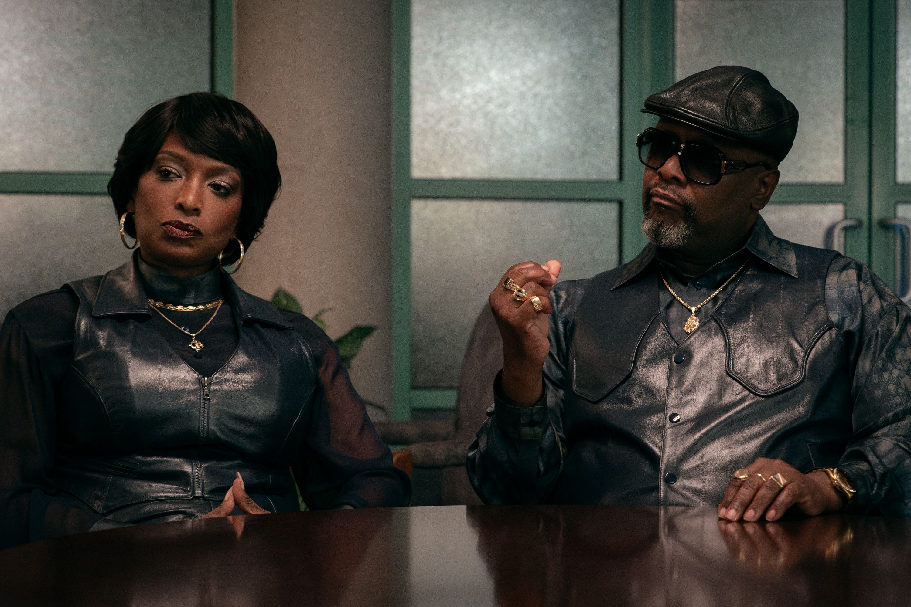Two well-dressed individuals, a woman and a man, sitting at a table and conversing. The man is wearing sunglasses and a leather jacket, while the woman has a short hairstyle and is wearing jewelry.