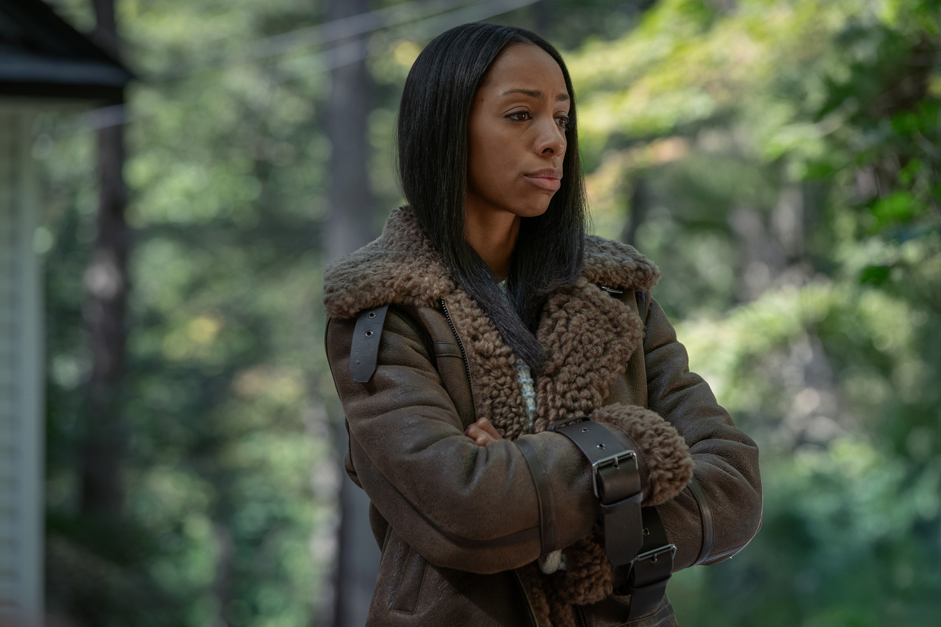 A woman in a brown fur-lined coat standing in a wooded area, looking thoughtful.