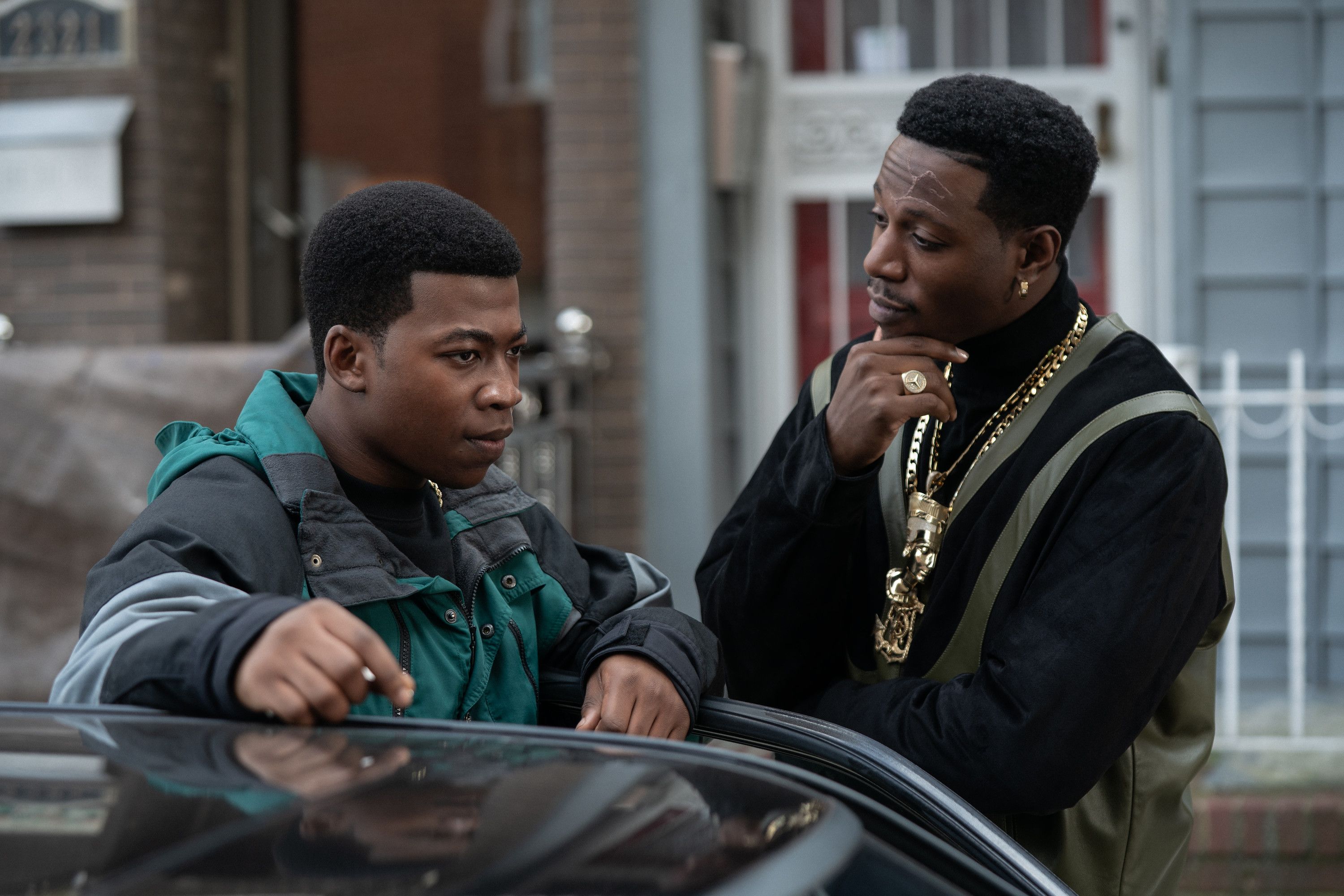 Two Black men conversing on a city street, one wearing a green jacket and the other wearing a black coat with gold jewelry.