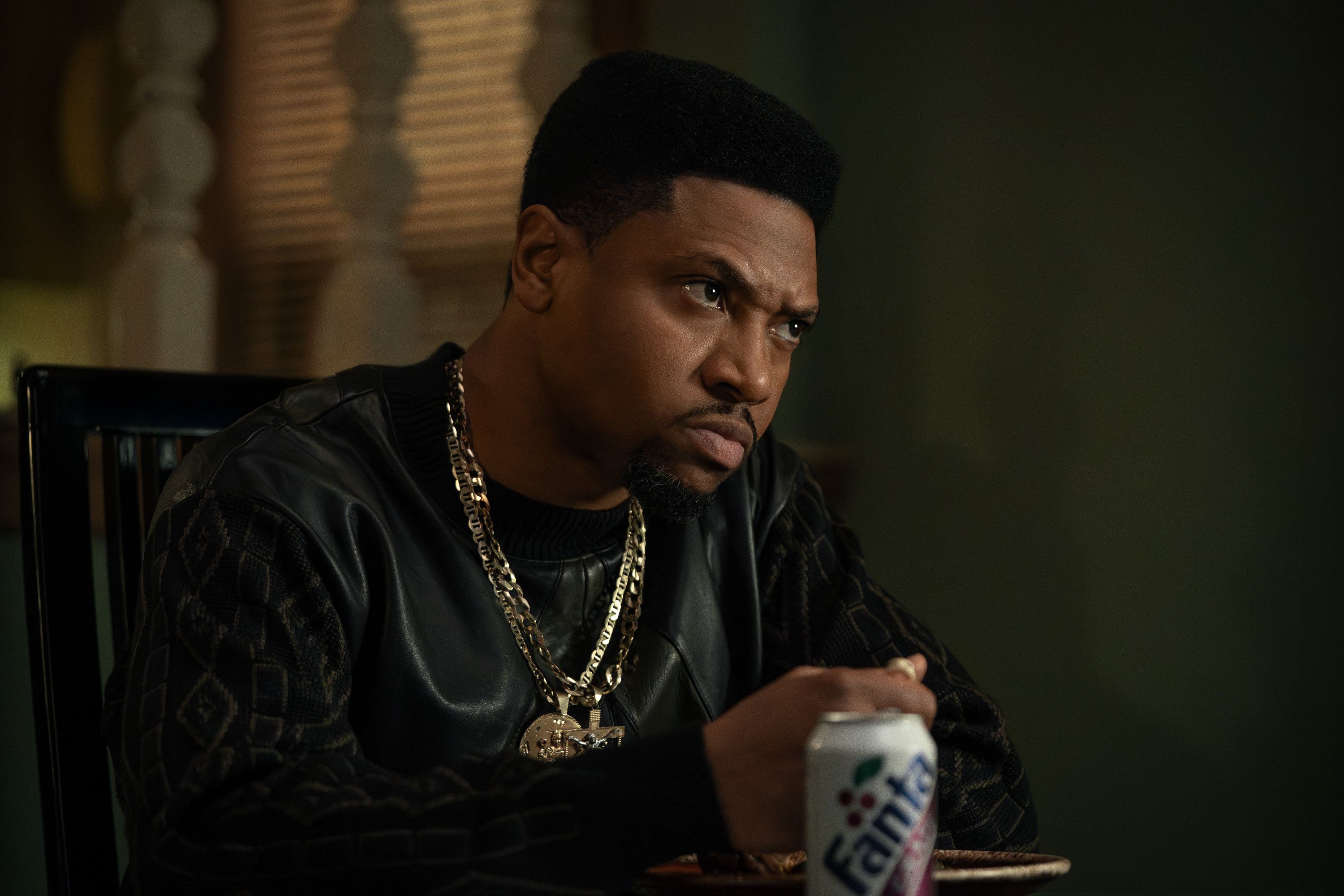 A serious-looking man wearing a black leather jacket and gold chains, holding a can of Sprite soda.