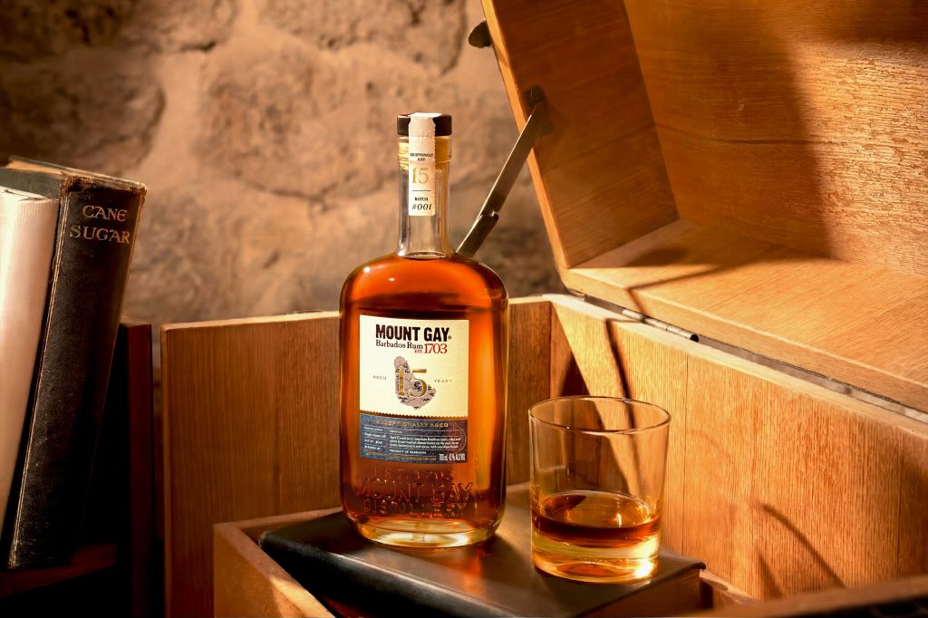 A bottle of Mount Gay rum and a glass of the amber-colored liquor on a wooden surface, surrounded by other wooden objects.