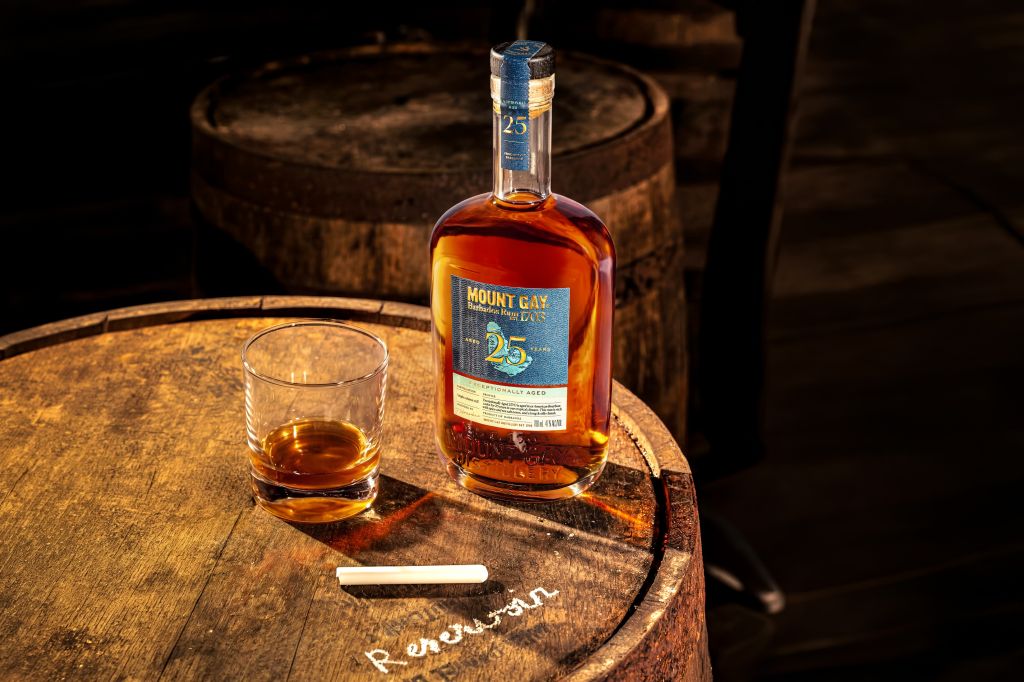 A bottle of Mount Gay 25-year-old rum on a wooden barrel, with a glass of the amber-colored liquor and a cigar beside it.