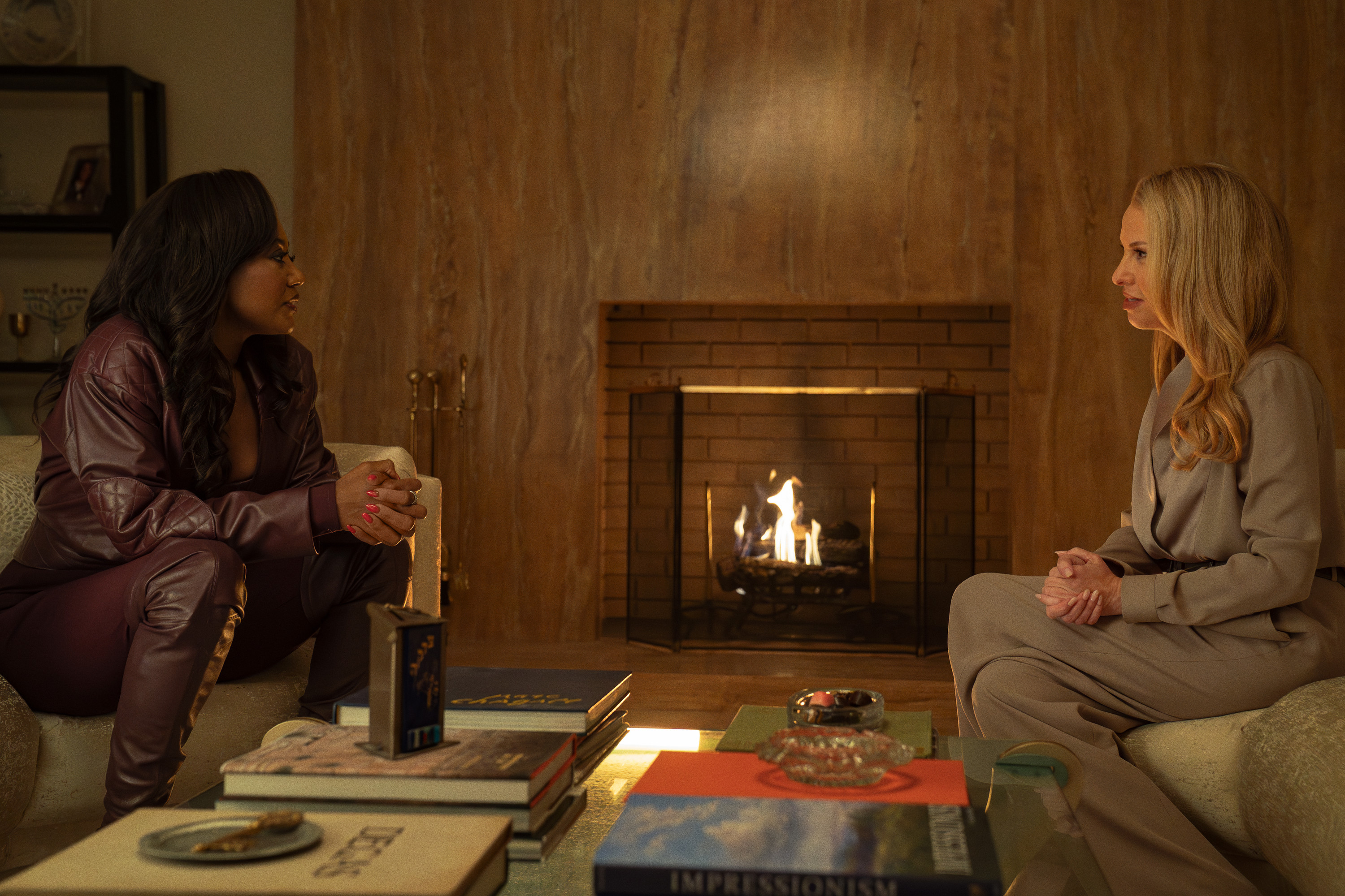 Two women sitting by a fireplace, one in a purple leather jacket and the other in a gray sweater, engaged in conversation.