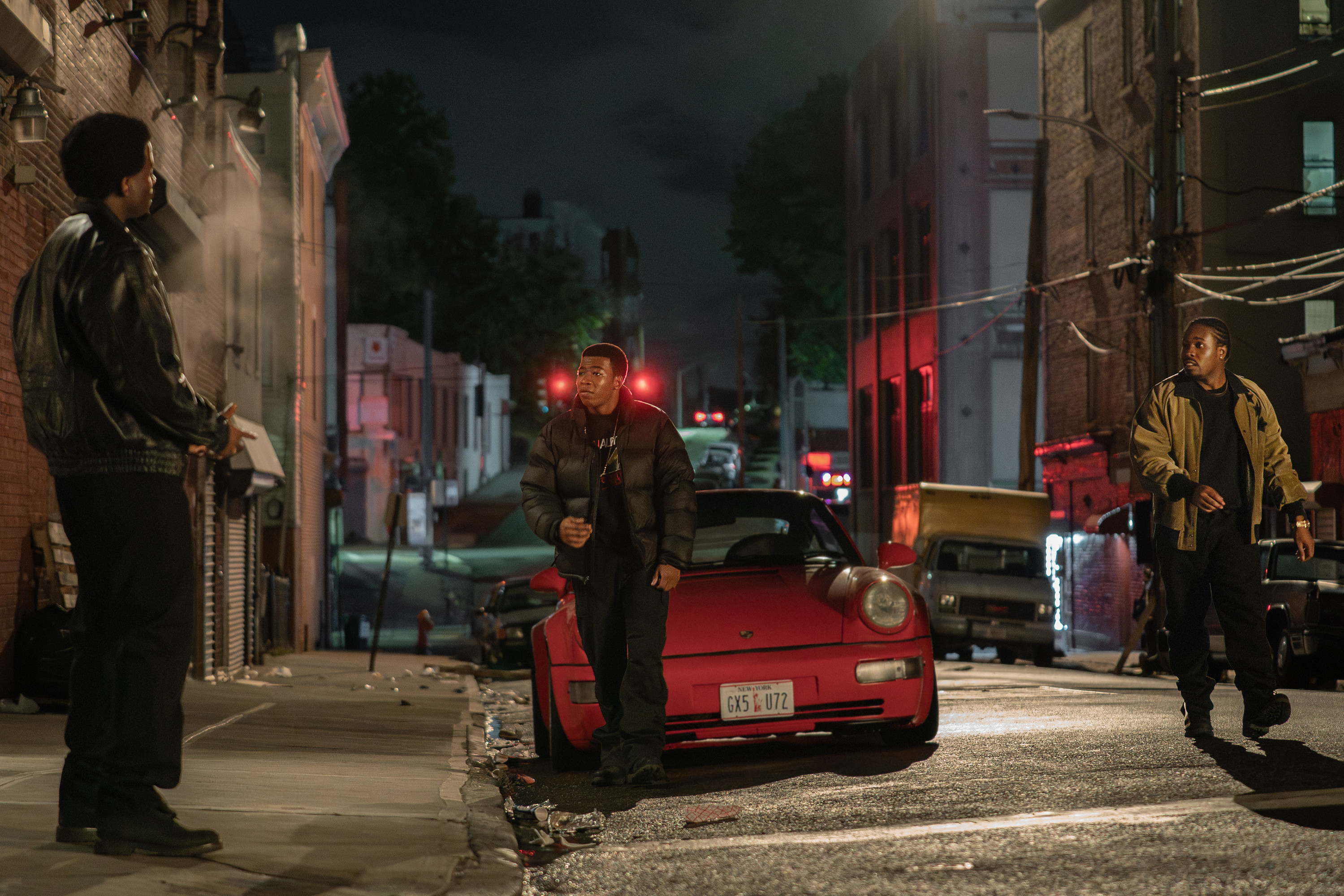 Three men standing on a dark city street at night, with a red sports car parked in the foreground.