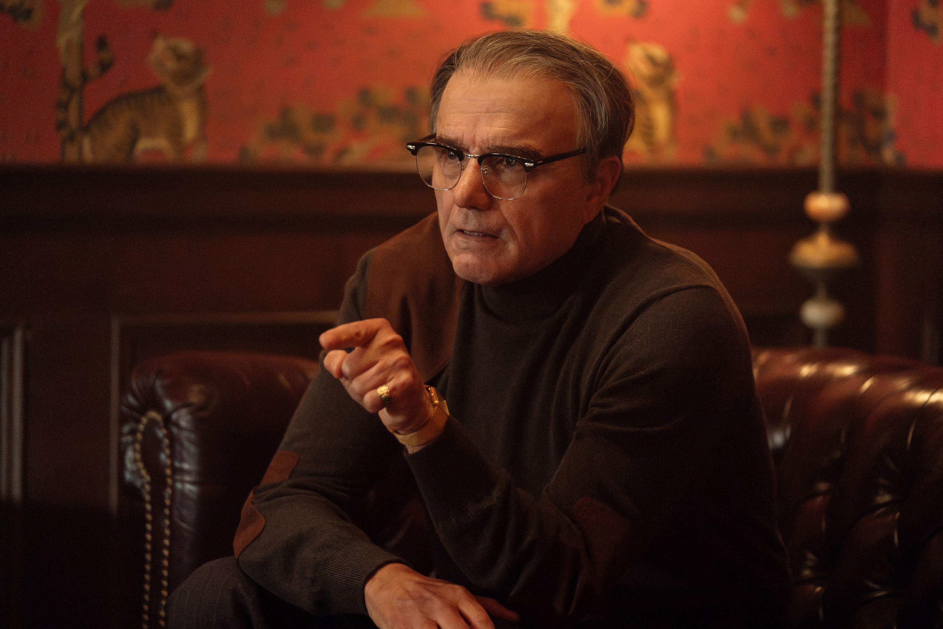 A middle-aged man with glasses and a serious expression, sitting in a dimly lit room with a red patterned wall behind him.