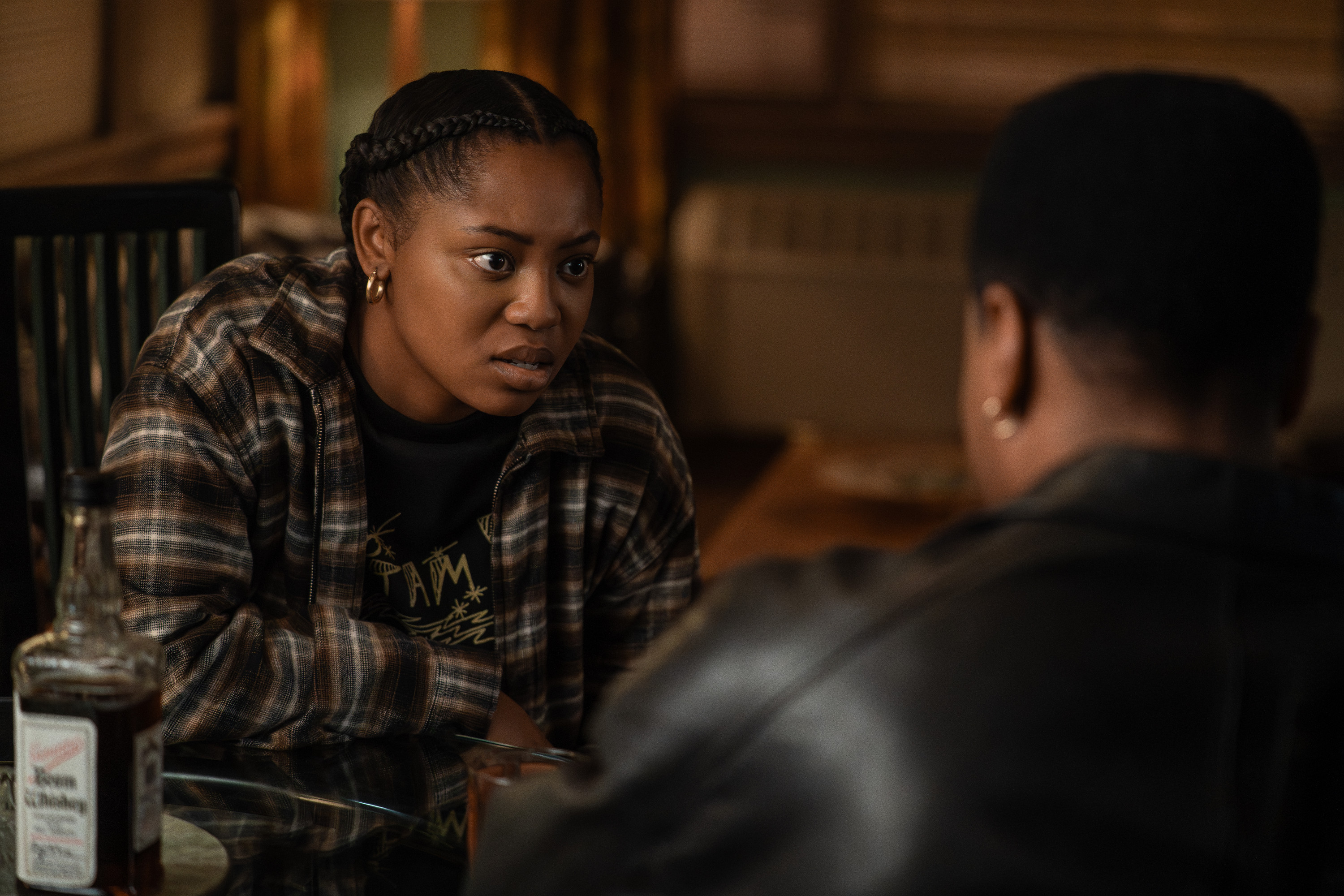 A young woman with braided hair wearing a plaid shirt and a t-shirt with text, sitting at a table and looking intently at someone out of frame.