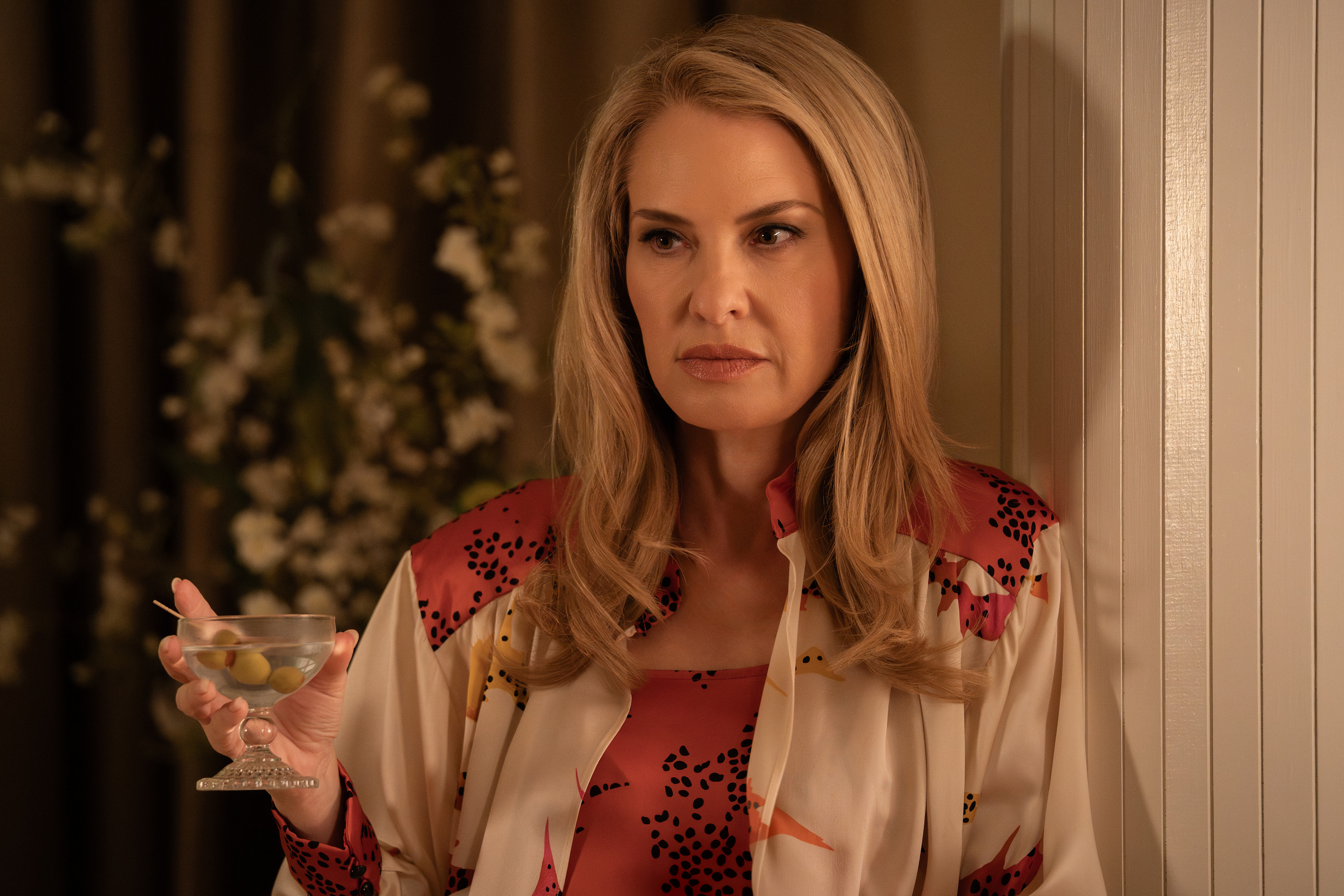 A woman with long blonde hair wearing a red and white patterned dress and holding a martini glass, standing in a dimly lit room with a wooden wall.