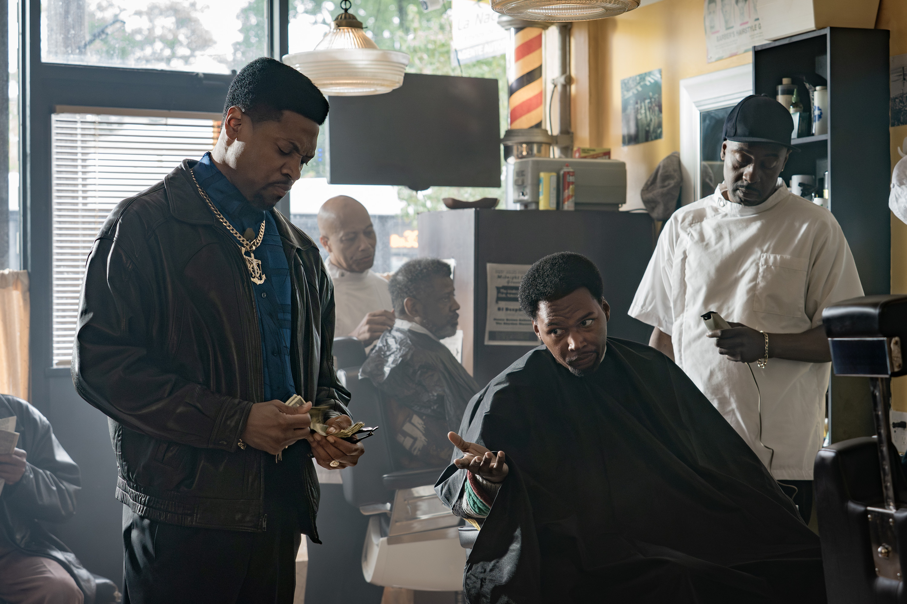 Three Black men in a barbershop, one wearing a gold chain and jacket, another getting a haircut, and a third standing in the background.