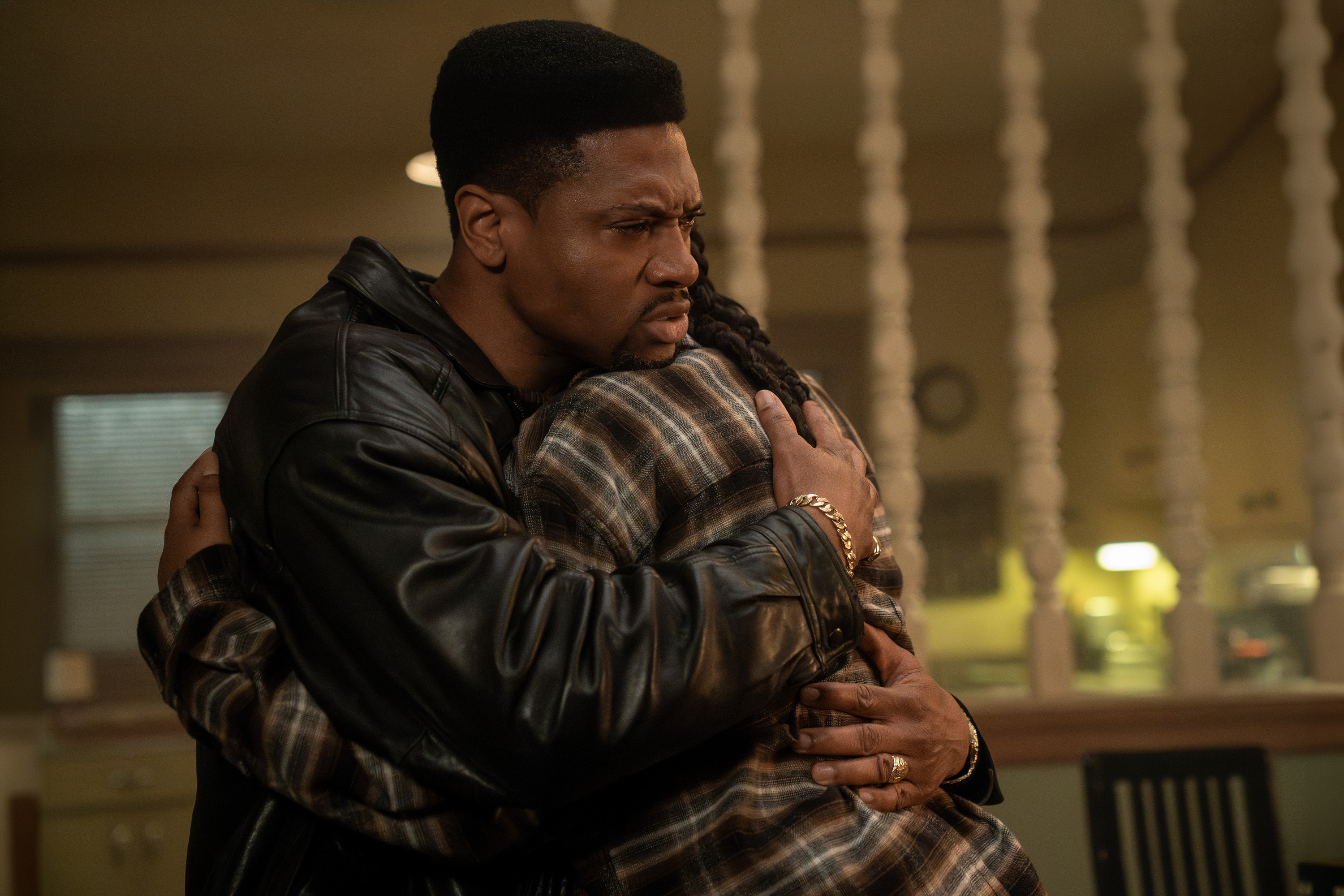 A man in a leather jacket embracing another person in a plaid shirt, with a pensive expression on his face.