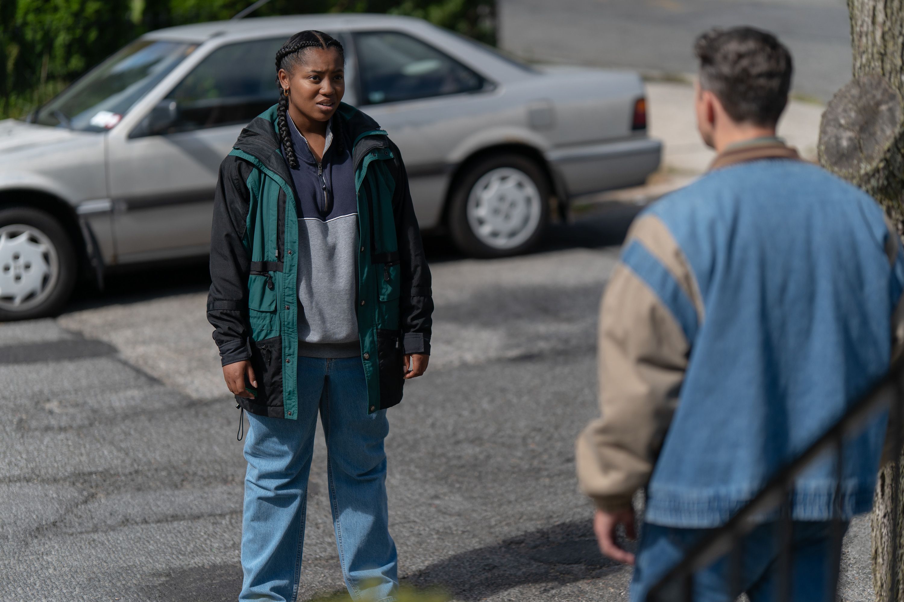 Two people, a woman in a green jacket and a man in a blue sweater, standing and conversing in a parking lot with a car visible in the background.