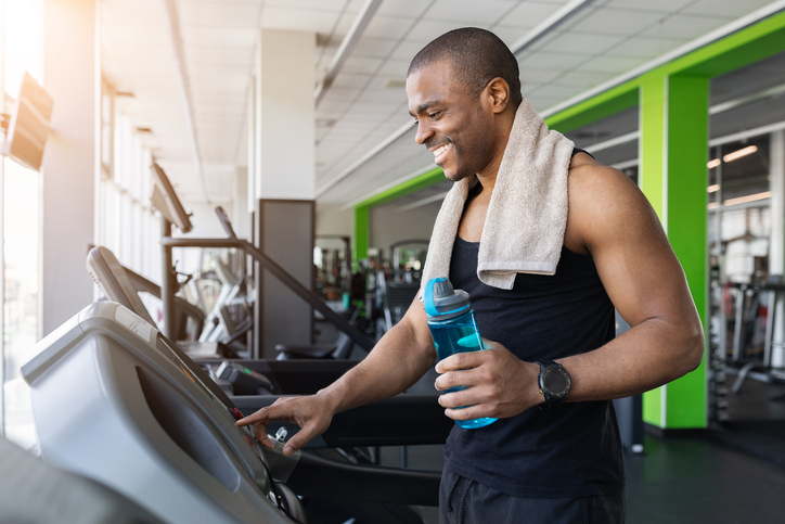 Young black man adjusting treadmill settings while holding water bottle in gym