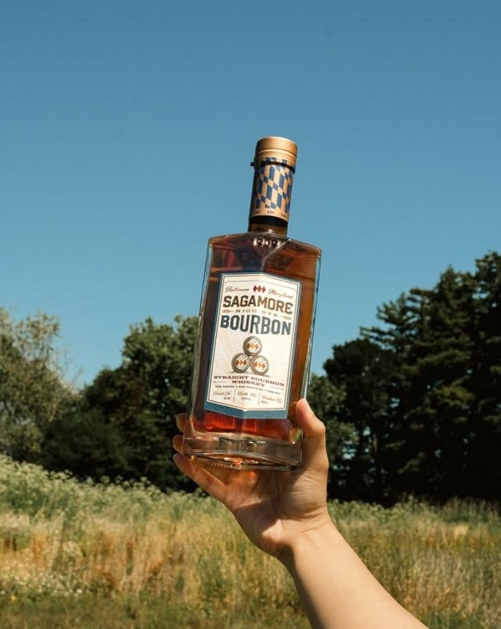 A hand holding a bottle of Sagamore Bourbon against a backdrop of trees and a blue sky.