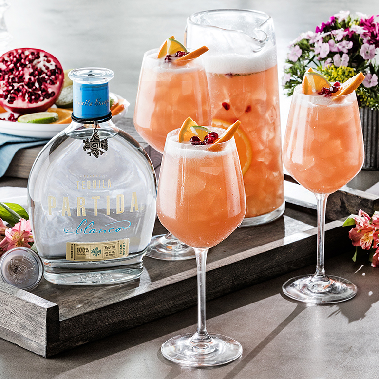 A tray with glasses of pink cocktail garnished with orange slices and pomegranate seeds, alongside a bottle of Milagro Partida tequila.