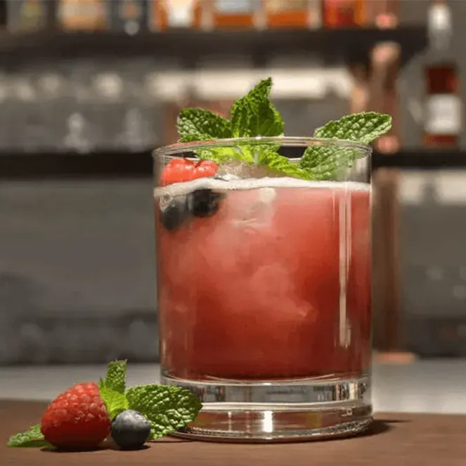 A glass of red cocktail garnished with mint leaves and fresh berries, set against a blurred background.