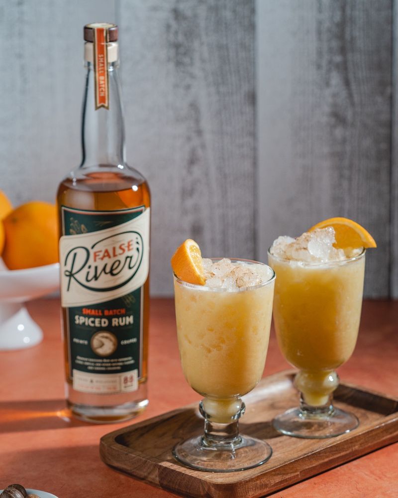 A bottle of False River spiced rum and two glasses filled with an orange-colored cocktail garnished with orange slices on a wooden board.