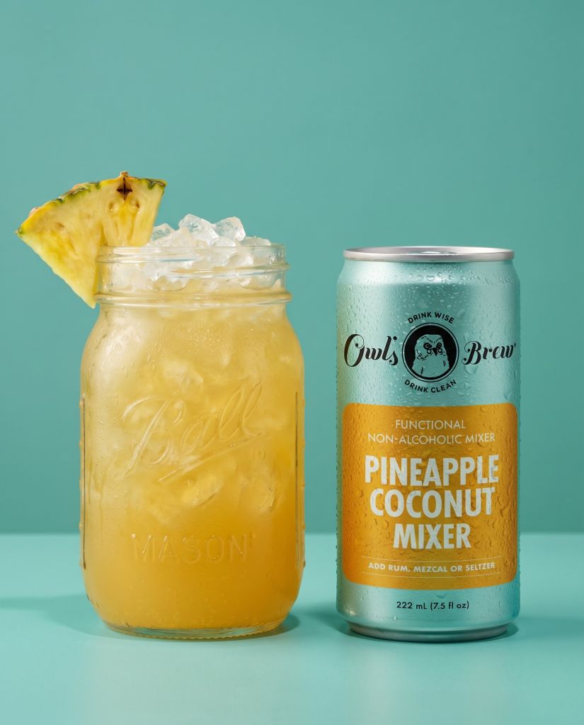 A glass mason jar filled with a pineapple and coconut-based non-alcoholic mixer, accompanied by a can of Owl's Brew Pineapple Coconut Mixer.
